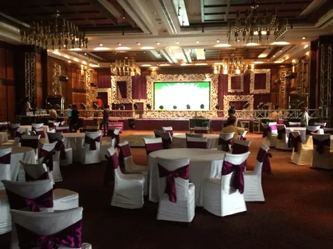 A banquet hall of round tables in plum sashes facing an ornate white stage and LED wall