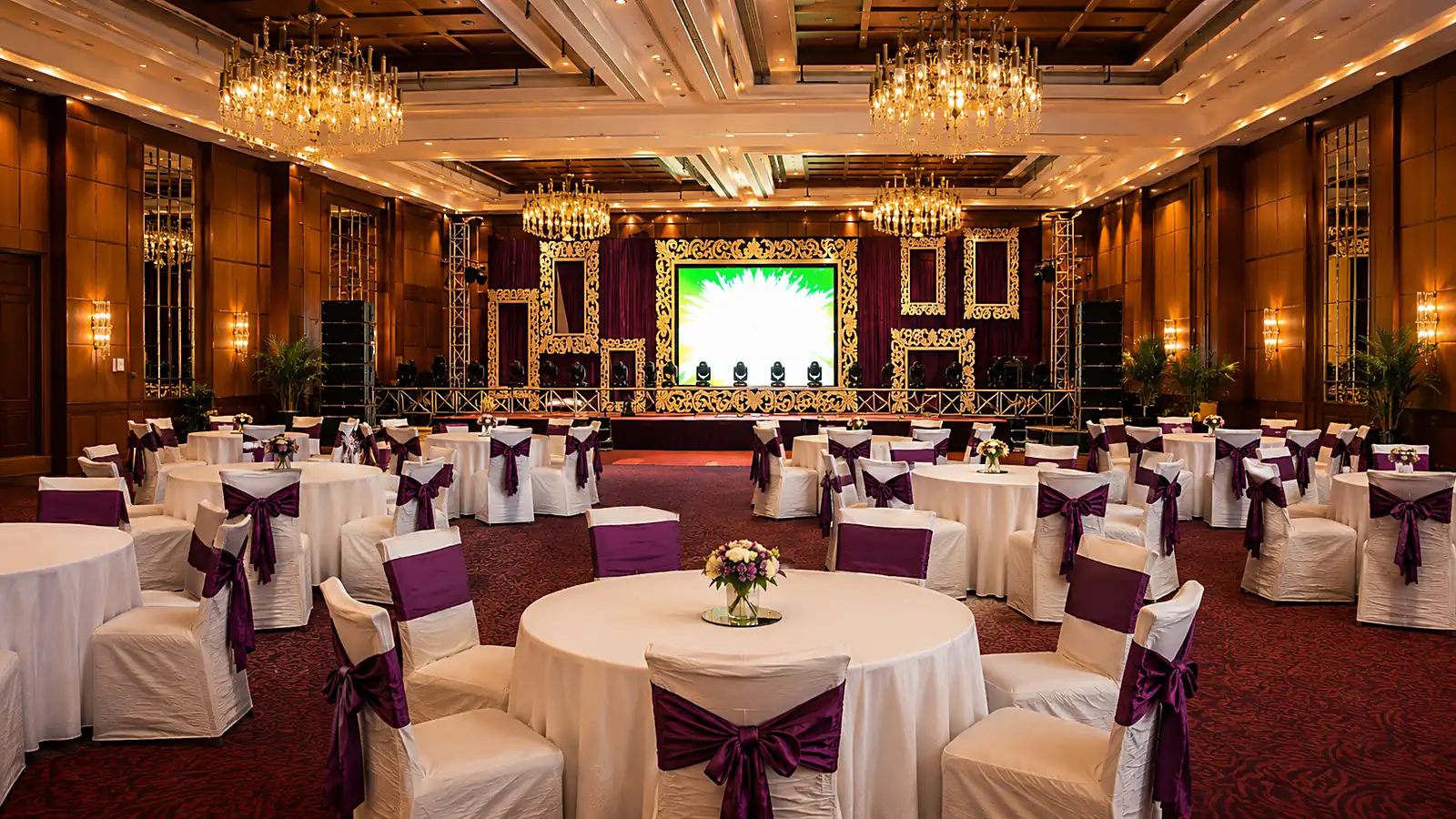 Banquet hall set for a corporate evening, chandeliers overhead and a lit gold stage beyond round tables dressed in plum sashes