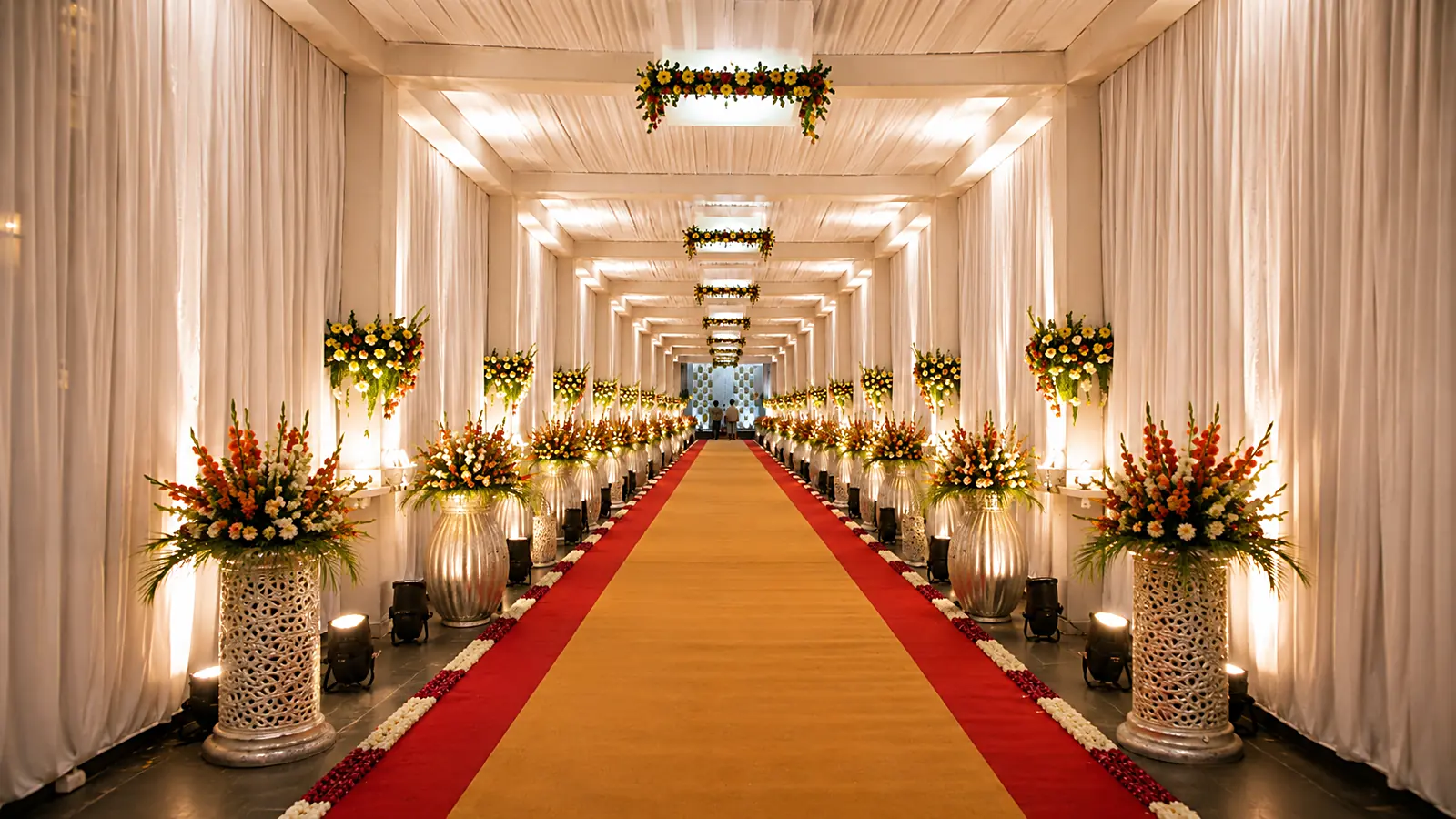 An ivory-draped entrance corridor with floral pillars either side of a red and gold aisle running to a lit archway