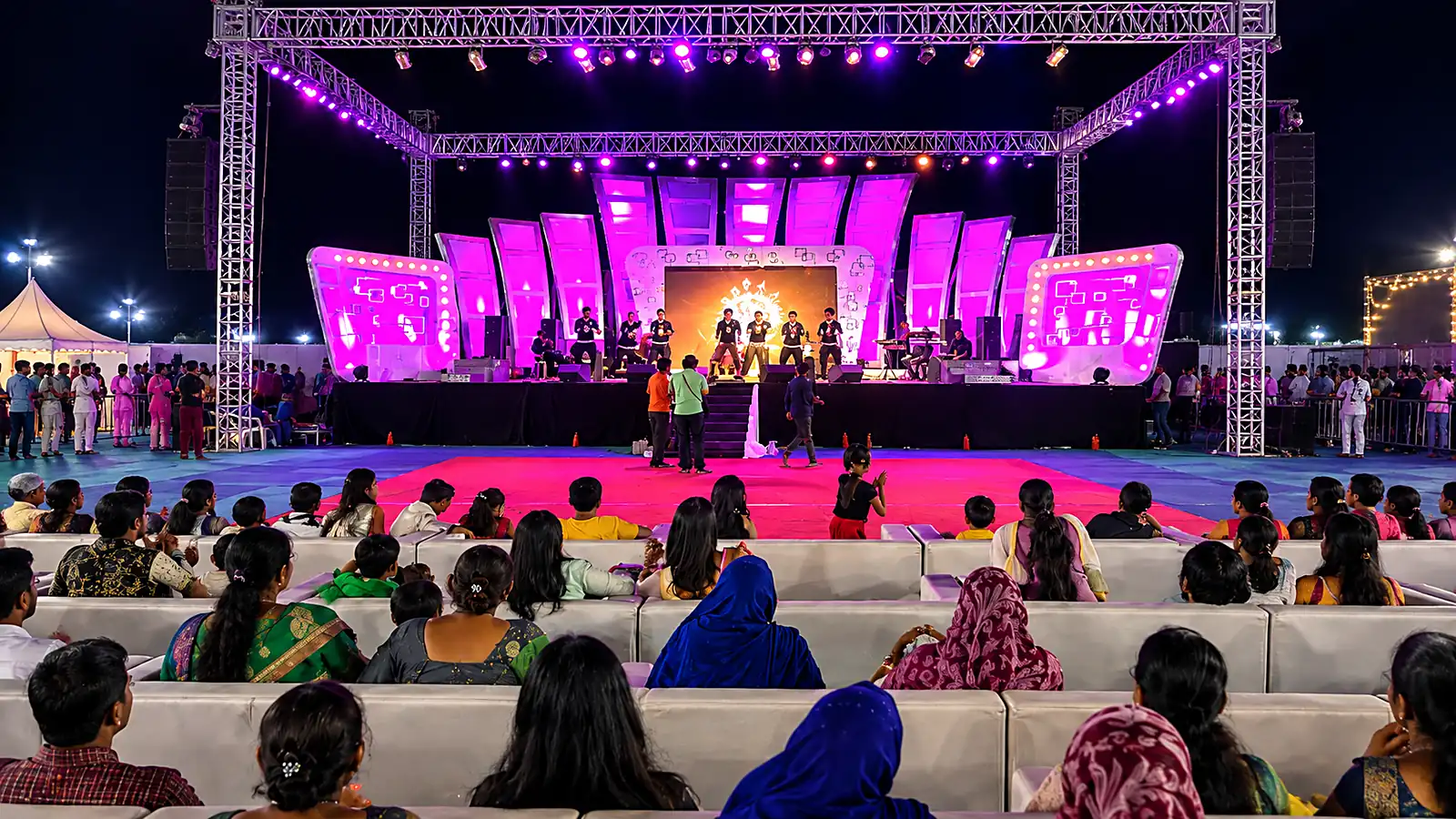 An outdoor stage washed in magenta under a lighting truss, performers on stage and a seated audience in front