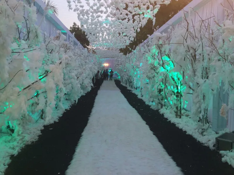A white snow-forest entrance tunnel lit in teal, its branches converging toward a lit archway